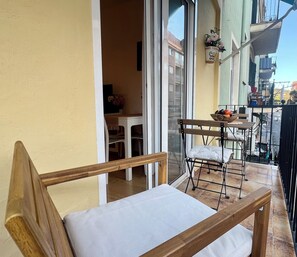 Un balcón con una silla de madera, una mesa pequeña con un frutero y una planta en maceta.