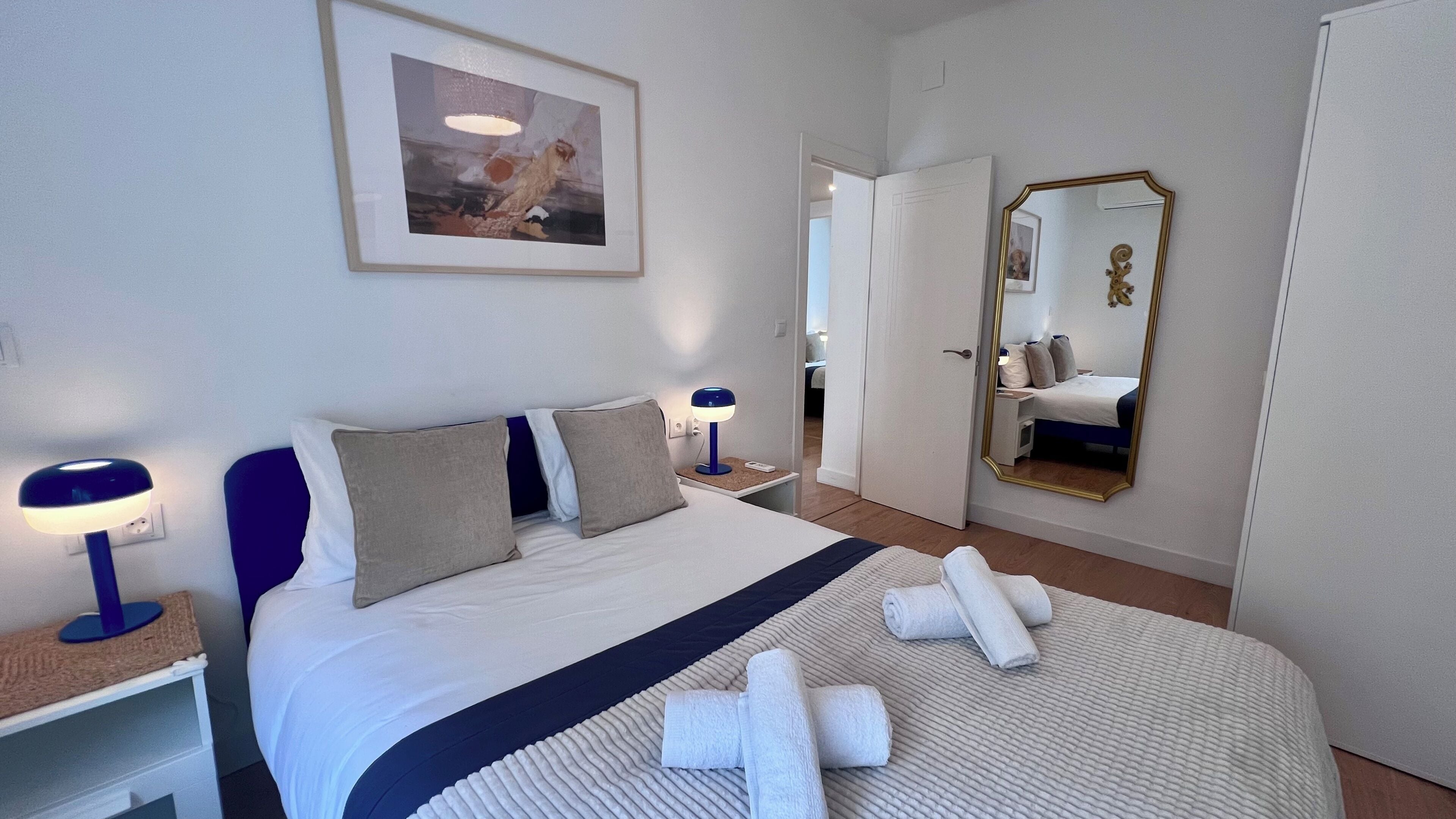 A neatly made bed with pillows, a bedside lamp, a framed picture on the wall, and a mirror on the adjacent wall.