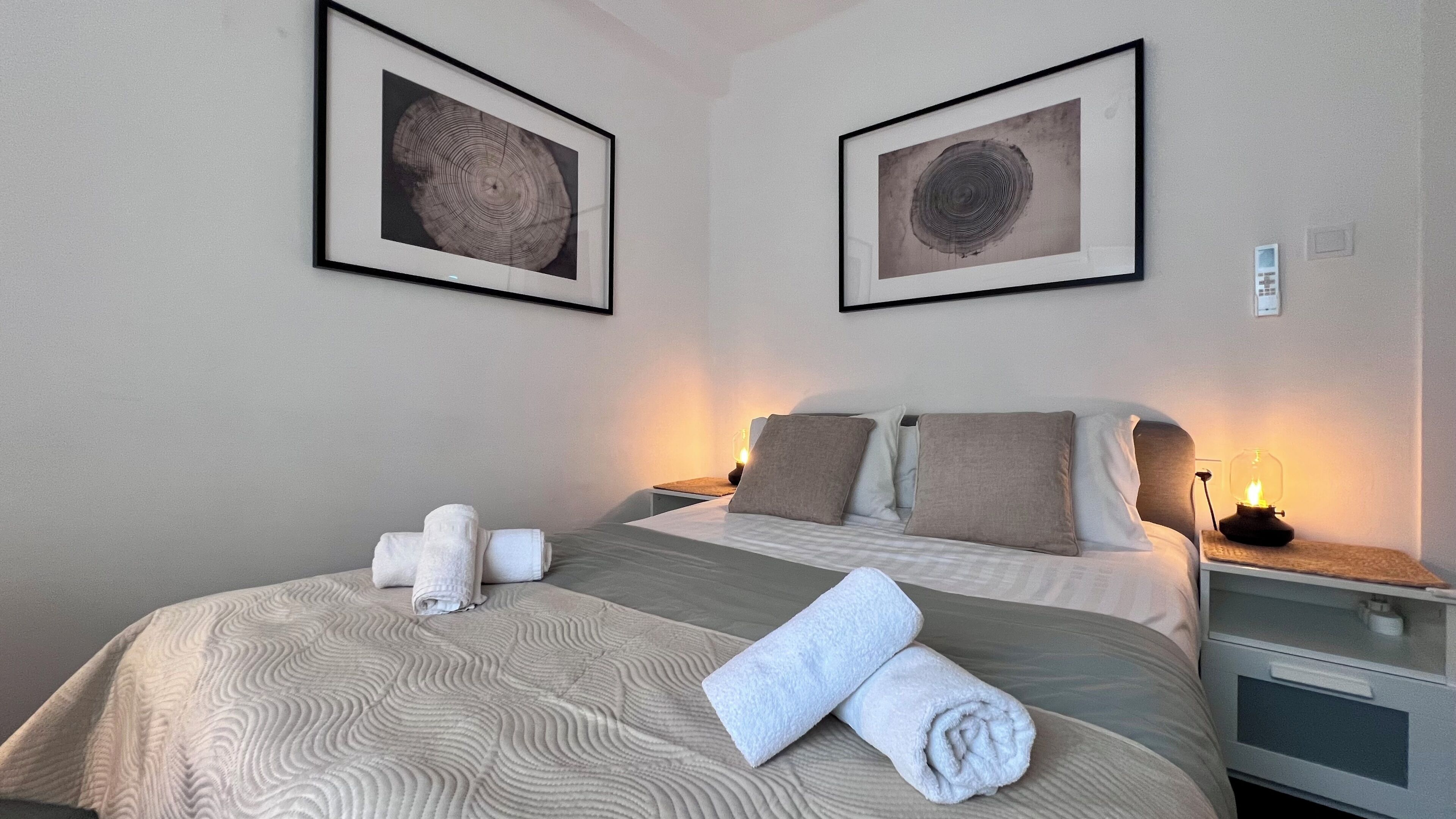 Ein Hotelzimmer mit einem Bett, zwei gerahmten Bildern an der Wand, einem Nachttisch mit einer Lampe und einem zusammengerollten Handtuch auf dem Bett.