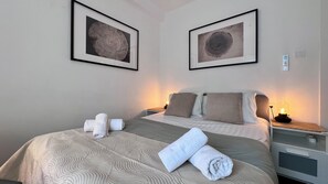 Habitación de hotel con cama, dos cuadros en la pared, una mesita de noche con lámpara y una toalla enrollada sobre la cama.