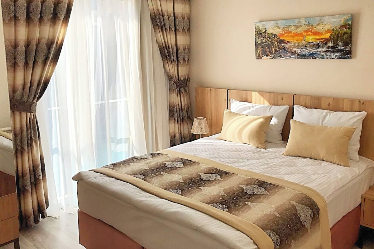 A bedroom with a bed, curtains, a painting, and a nightstand.