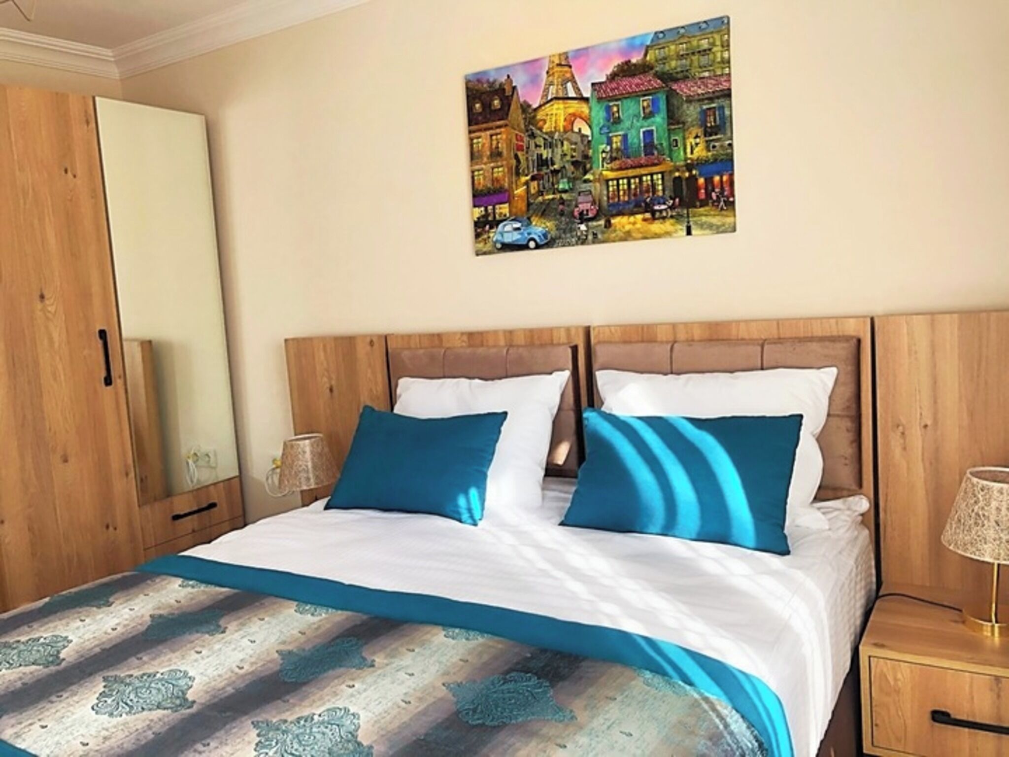 Quarto com cabeceira de madeira, cama com roupa de cama branca e azul, mesa de cabeceira com abajur e um quadro de uma paisagem urbana na parede.