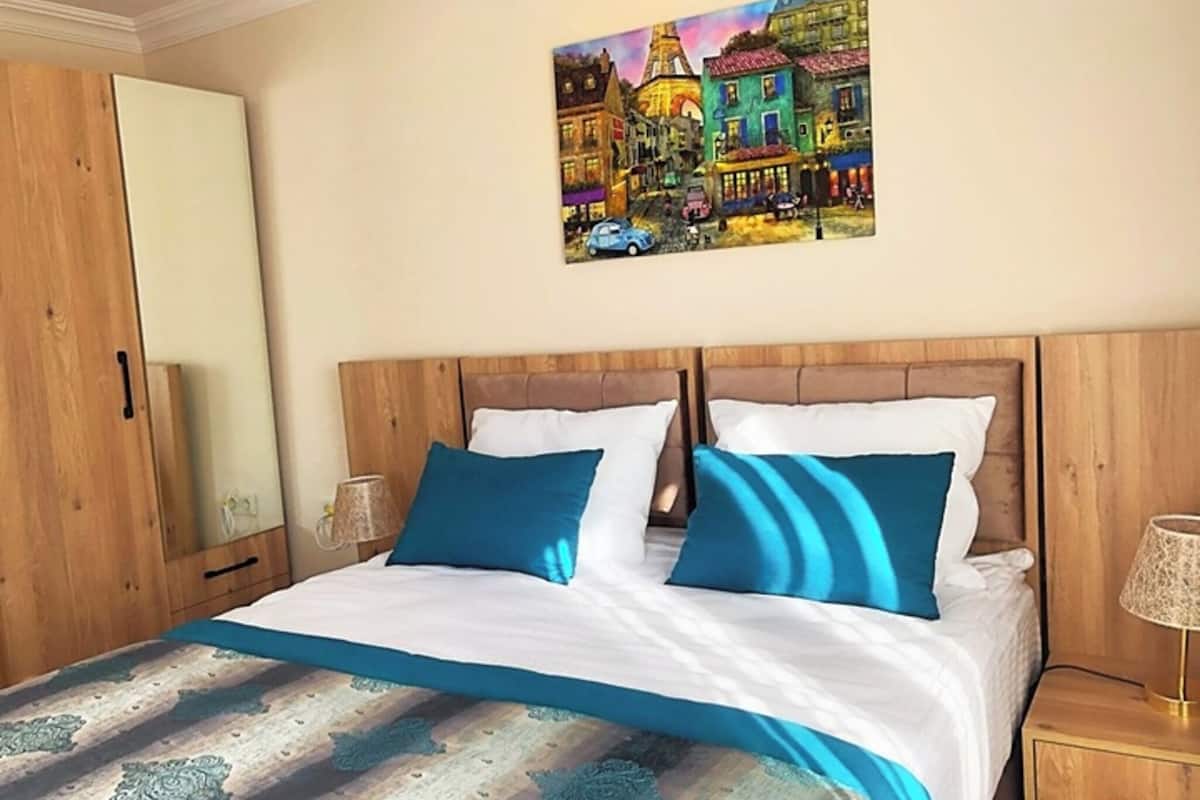 A bedroom with a wooden headboard, a bed with white and blue bedding, a nightstand with a lamp, and a painting of a cityscape on the wall.