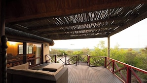 Una terraza de madera con barandilla roja, un sofá de mimbre y un ventanal que da a un espacio interior con vistas al paisaje.