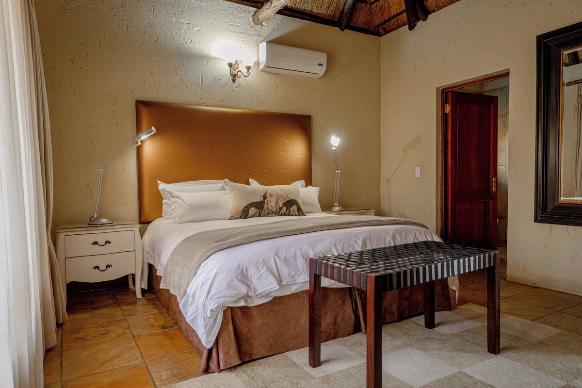 A bedroom with a wooden headboard, a bed with white bedding, a bedside table with a lamp, and a small wooden bench.