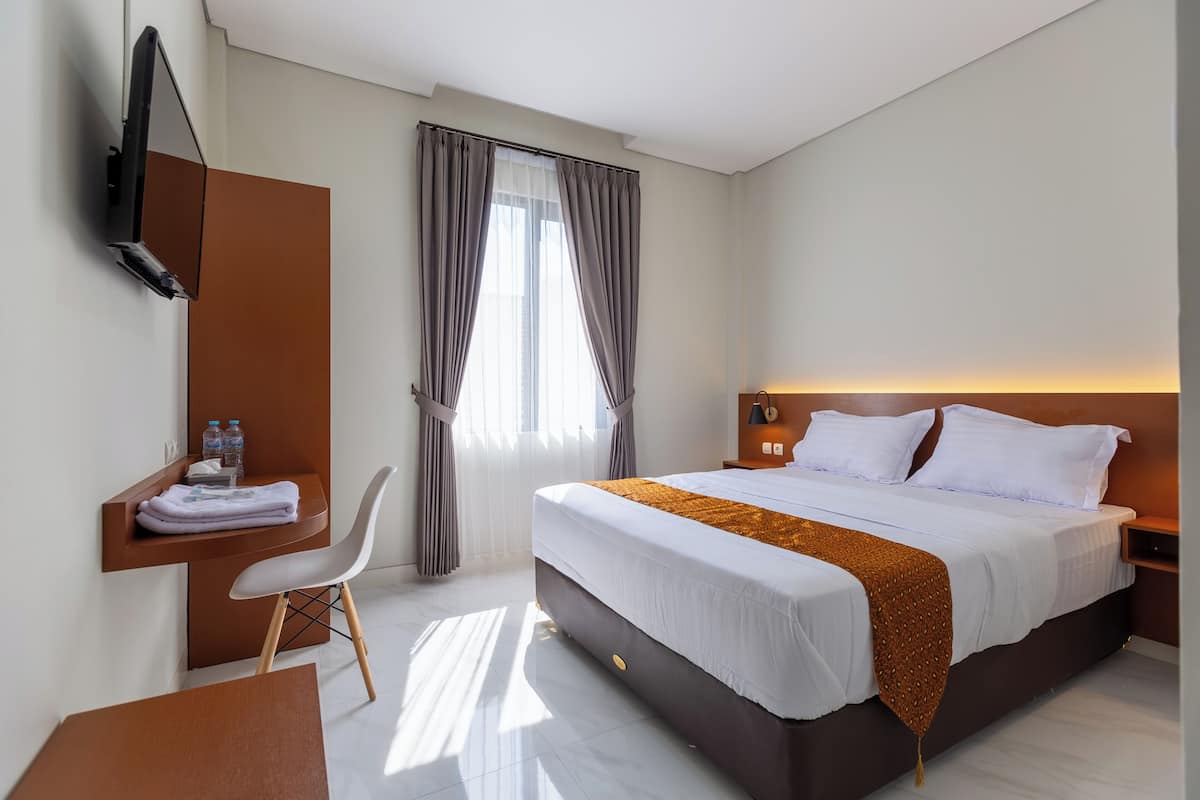 A modern hotel room with a bed, a desk with a chair, a TV, and a window with curtains.