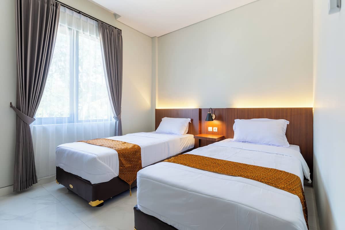 A hotel room with two beds, a window with curtains, a nightstand with a lamp, and a wooden headboard.