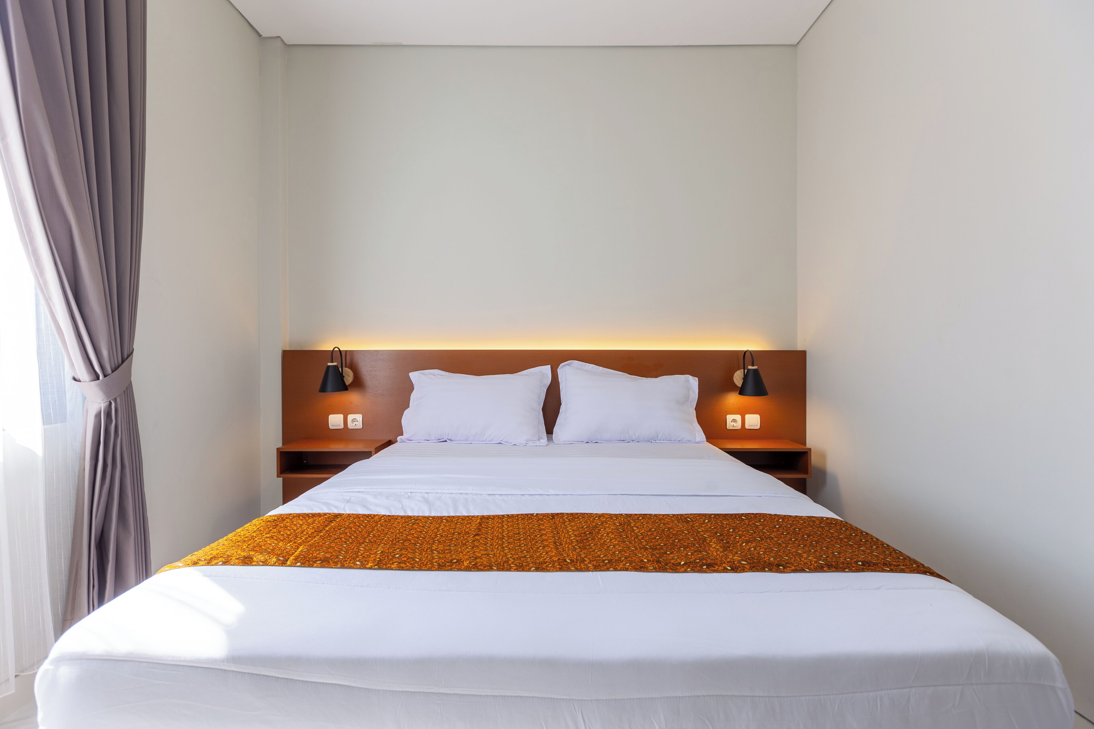 Una cama bien tendida con sábanas blancas y una manta marrón, dos lámparas de noche y un cabecero de madera.