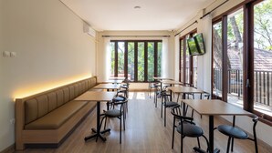 Un comedor moderno con un banco largo, mesas y sillas, ventanas amplias con cortinas y un televisor de pantalla plana montado en la pared.