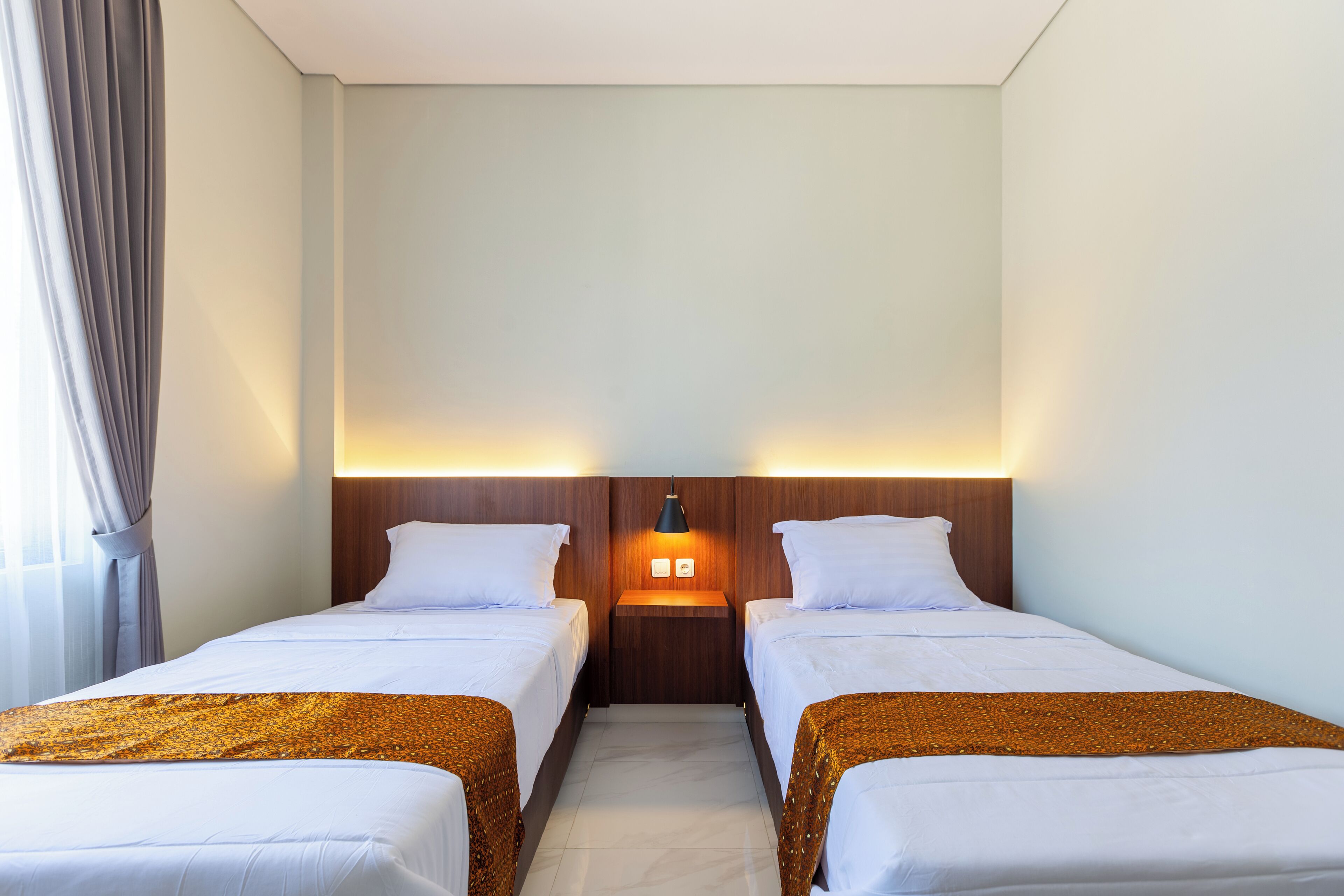 Hotellihuone, jossa on kaksi erillistä sänkyä, yöpöytä lampun kanssa ja ikkuna, jossa on verhot.