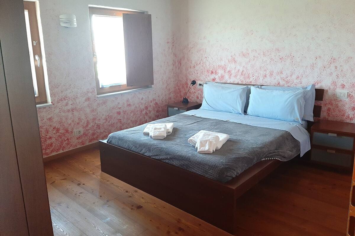A bedroom with a wooden bed, a nightstand, a window, and a wall with floral wallpaper.