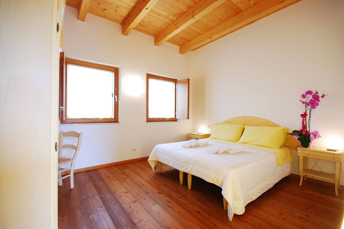 A bedroom with a wooden floor, a bed with yellow bedding, two windows, a chair, and a nightstand with a lamp.