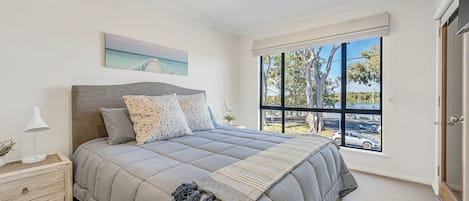 Una camera da letto con un letto, un comodino, una lampada, una finestra vista alberi e strada, e una ventola a soffitto.