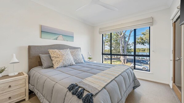 A bedroom with a bed, a nightstand, a lamp, a window with a view of trees and a road, and a ceiling fan.