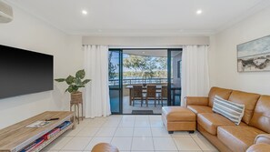 A modern living room with a flat-screen TV, a leather sofa, a dining area with wicker chairs, and a view of the outdoors.