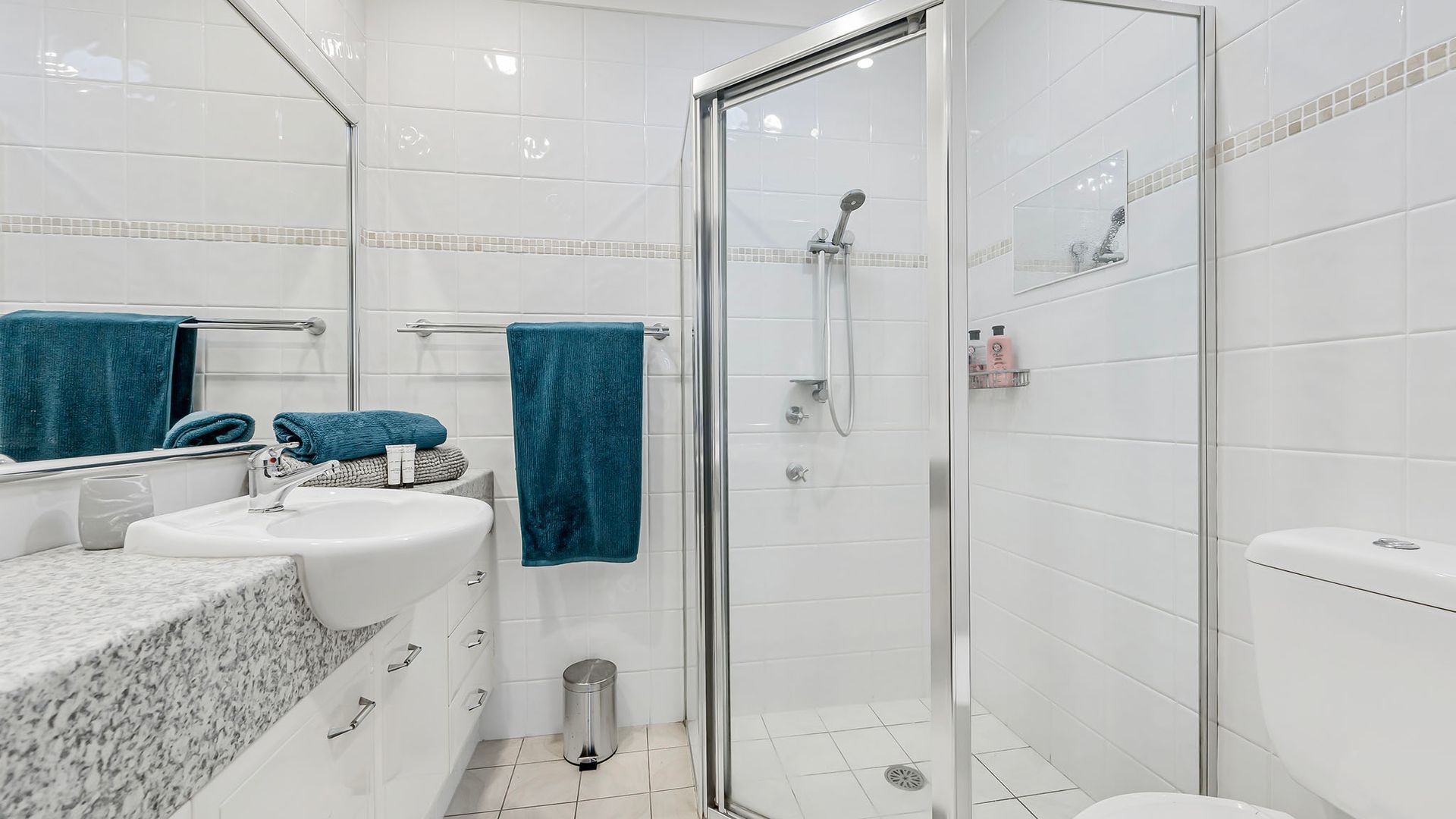 Casa de banho moderna com chuveiro envidraçado, azulejos brancos, bancada de mármore e sanita.