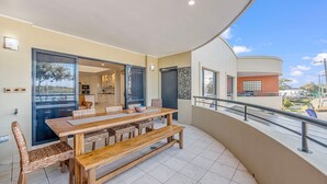 Uma sala de jantar com mesa de madeira e cadeiras de vime, cozinha visível por uma porta aberta, e varanda com guarda-corpo com vista para uma área residencial.