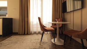 A modern hotel room with a dining area featuring a round table and two chairs, a flat-screen TV mounted on the wall, and a large window with sheer curtains. - Nest Inn Charm Apartment (Baia Mare)