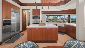 A modern kitchen with a large island, stainless steel appliances, wooden cabinetry, and a view of the outdoors. - Waiulu Estate 152B – Ocean View & Fully Renovated | Onsite Staff & Daily Housekeeping (Kailua Kona)
