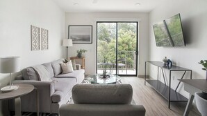 A modern living room with a grey sofa, a glass coffee table, and a balcony with outdoor furniture. - Landing | Spectacular 1BD, Courtyard, Gym (Austin)