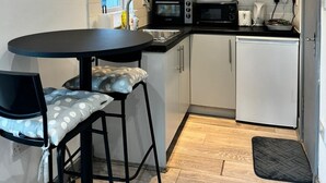 Cocina compacta con armarios blancos, encimera negra, microondas, tostadora y un refrigerador pequeño. Hay una mesa redonda con dos taburetes y una silla negra.