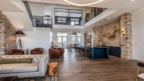 A spacious living area with a high ceiling, stone walls, a central seating area with sofas, a bar, and a staircase leading to an upper level. - Landing | Beautiful 1BD, Yoga Studio, Gym (Fort Worth)