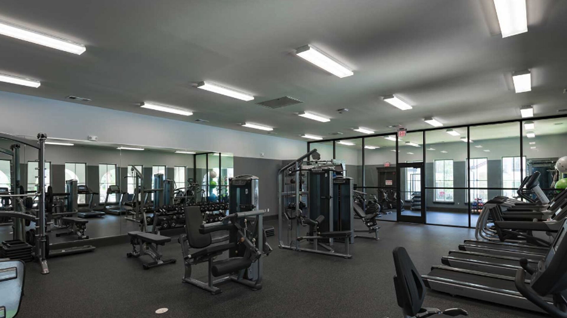 Un gimnasio con diversos equipos de ejercicio, paredes espejadas y grandes ventanales.