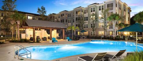 A hotel pool area with a clear blue pool, lounge chairs, and a covered seating area with tables and chairs.