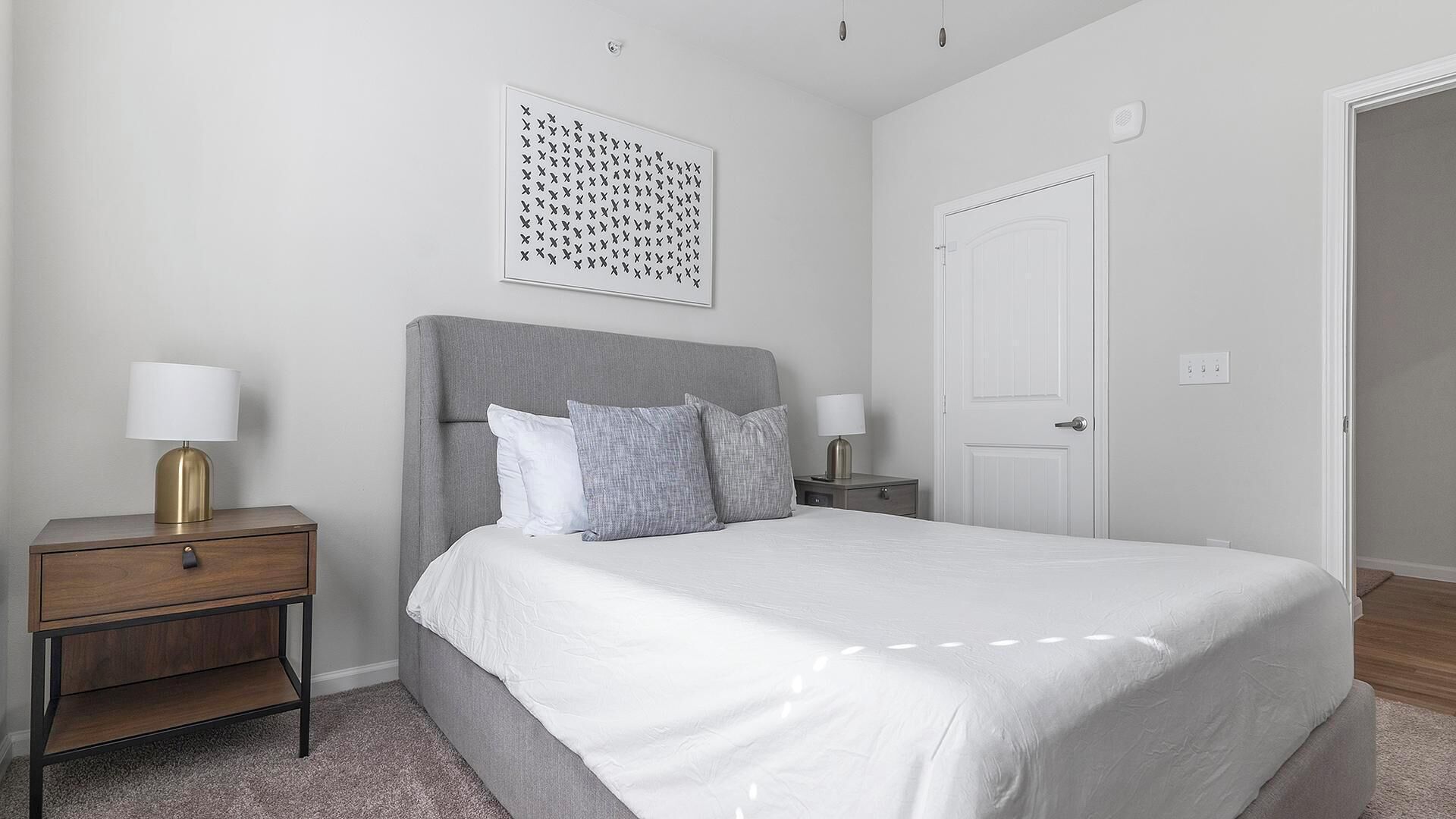A bedroom with a bed, two bedside tables, a nightstand, a white door, and a wall-mounted artwork.