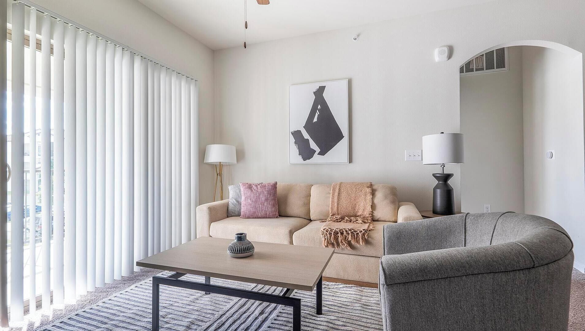 A modern living room with a sofa, a coffee table, a lamp, and a large window with blinds.