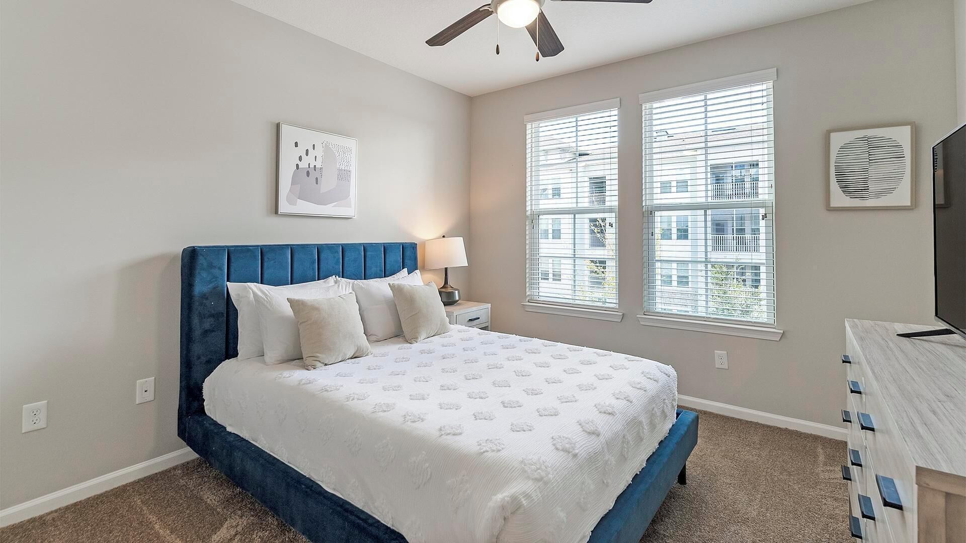 A bedroom with a bed, a nightstand, a lamp, a ceiling fan, and two windows with blinds.