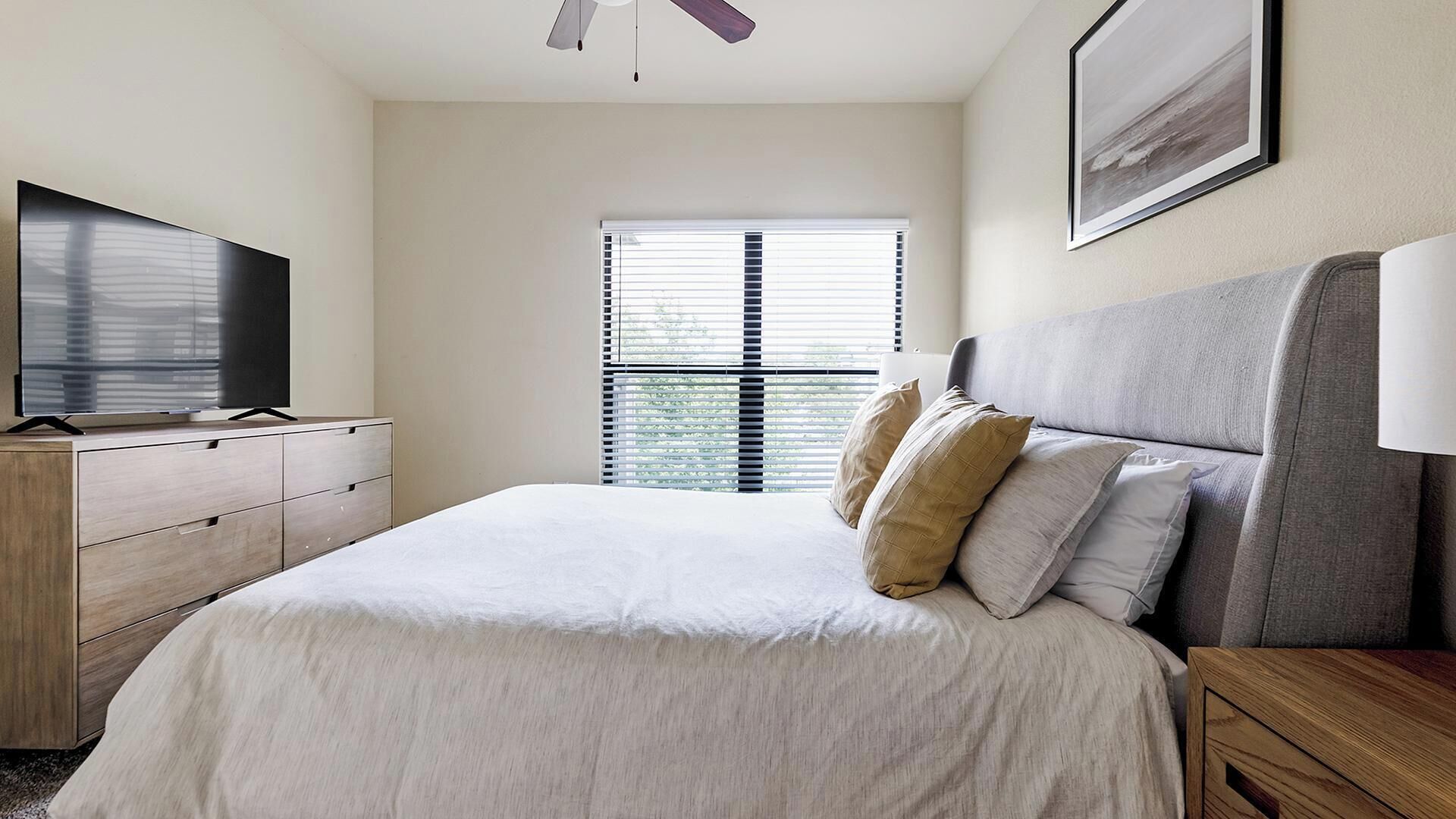 Une chambre d’hôtel avec un lit, une télévision, un ventilateur de plafond, une fenêtre avec des stores et un tableau au mur.