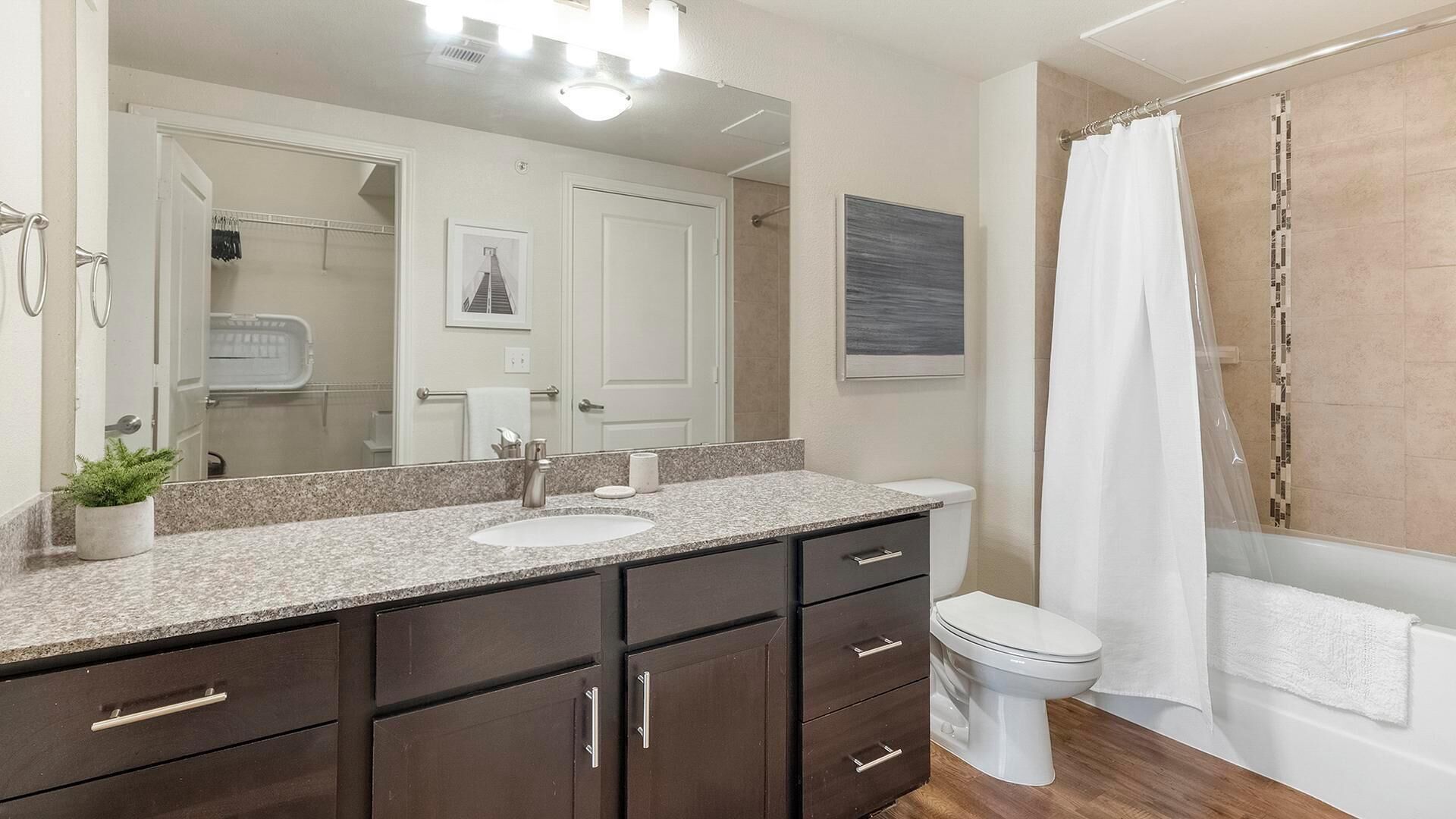 Un baño con un espejo grande, un lavabo, armarios de madera oscura, un inodoro y una ducha con cortina blanca.