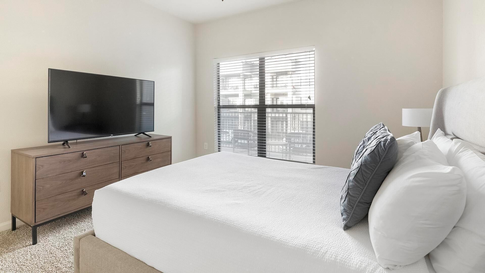 Une chambre à coucher avec un lit, un téléviseur posé sur une commode en bois et une fenêtre avec des stores.