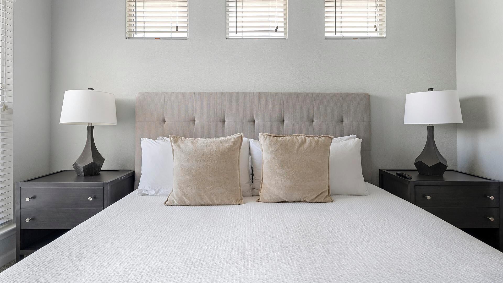 A bedroom with a large bed, two bedside tables with lamps, and three windows with blinds.