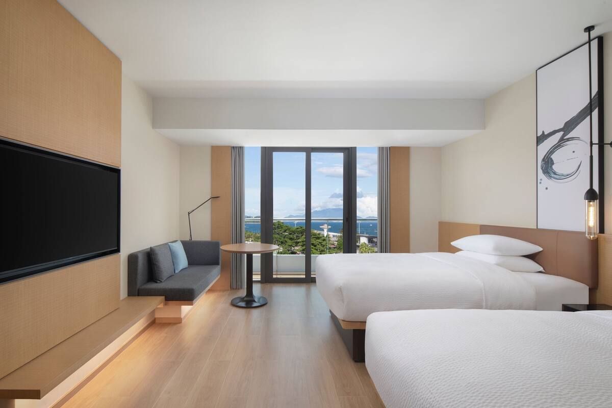 A modern hotel room with a large window, a sofa, two beds, and a flat-screen TV.