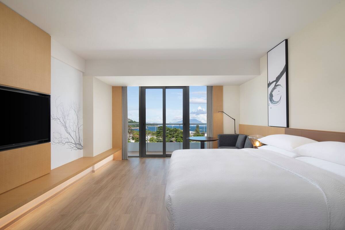 A modern hotel room with a large bed, a flat-screen TV, a desk with a chair, and a balcony with a view of the sea.