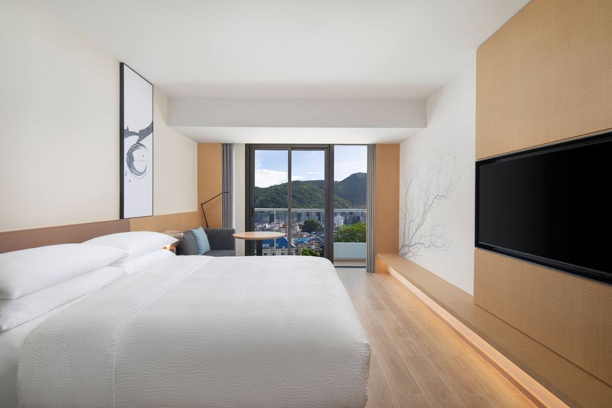 A modern hotel room with a large bed, a flat-screen TV, a small table, and a view of the city and mountains.