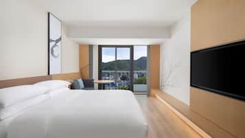 A modern hotel room with a large bed, a flat-screen TV, a small table, and a view of the city and mountains.