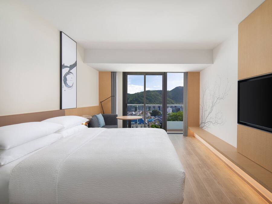 A modern hotel room with a large bed, a flat-screen TV, a small table, and a view of the city and mountains.