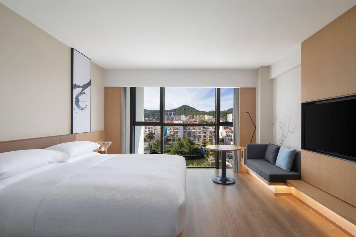 A modern hotel room with a large bed, a sofa, a TV, and a view of the cityscape.