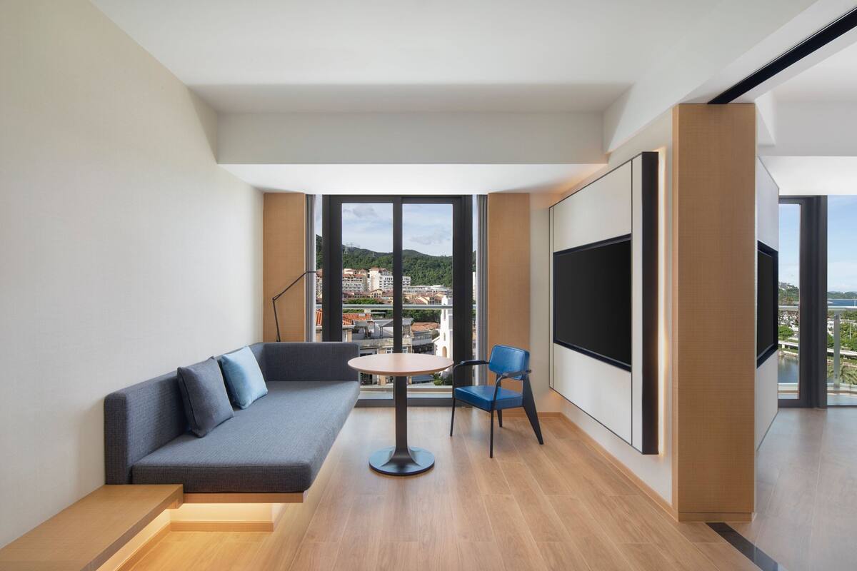 A modern living room with a sofa, a round table, and a chair, large windows offering a city view.