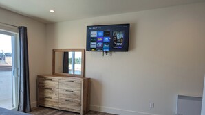 A bedroom with a large bed, a wooden dresser, a flat-screen TV mounted on the wall, and a window with curtains. - Downtown Friday Harbor! Newly Built! PM (Friday Harbor)