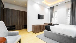 A modern hotel room with a grey sofa, a bed with white bedding, a TV mounted on the wall, and a wooden wardrobe. - Aaron's Place (Enugu)