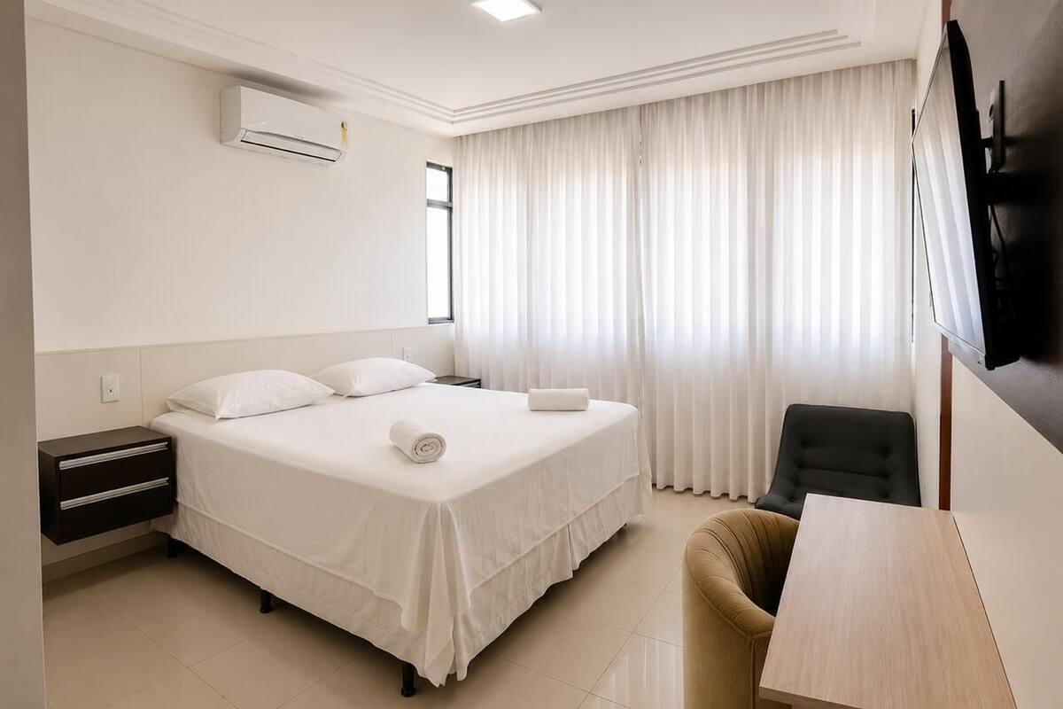 A hotel room with a bed, a desk, a chair, a TV, and an air conditioning unit.