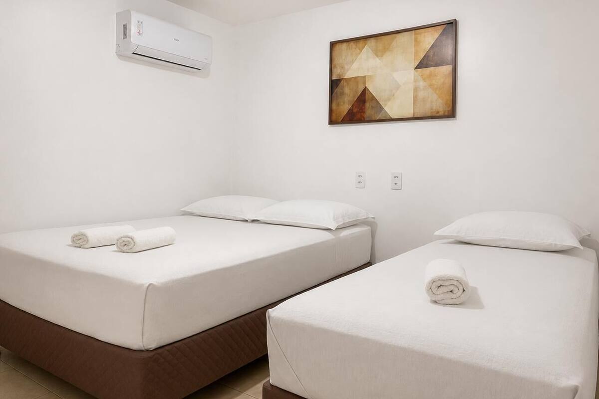 A hotel room with two beds, white linens, a wall-mounted air conditioner, and a geometric painting.