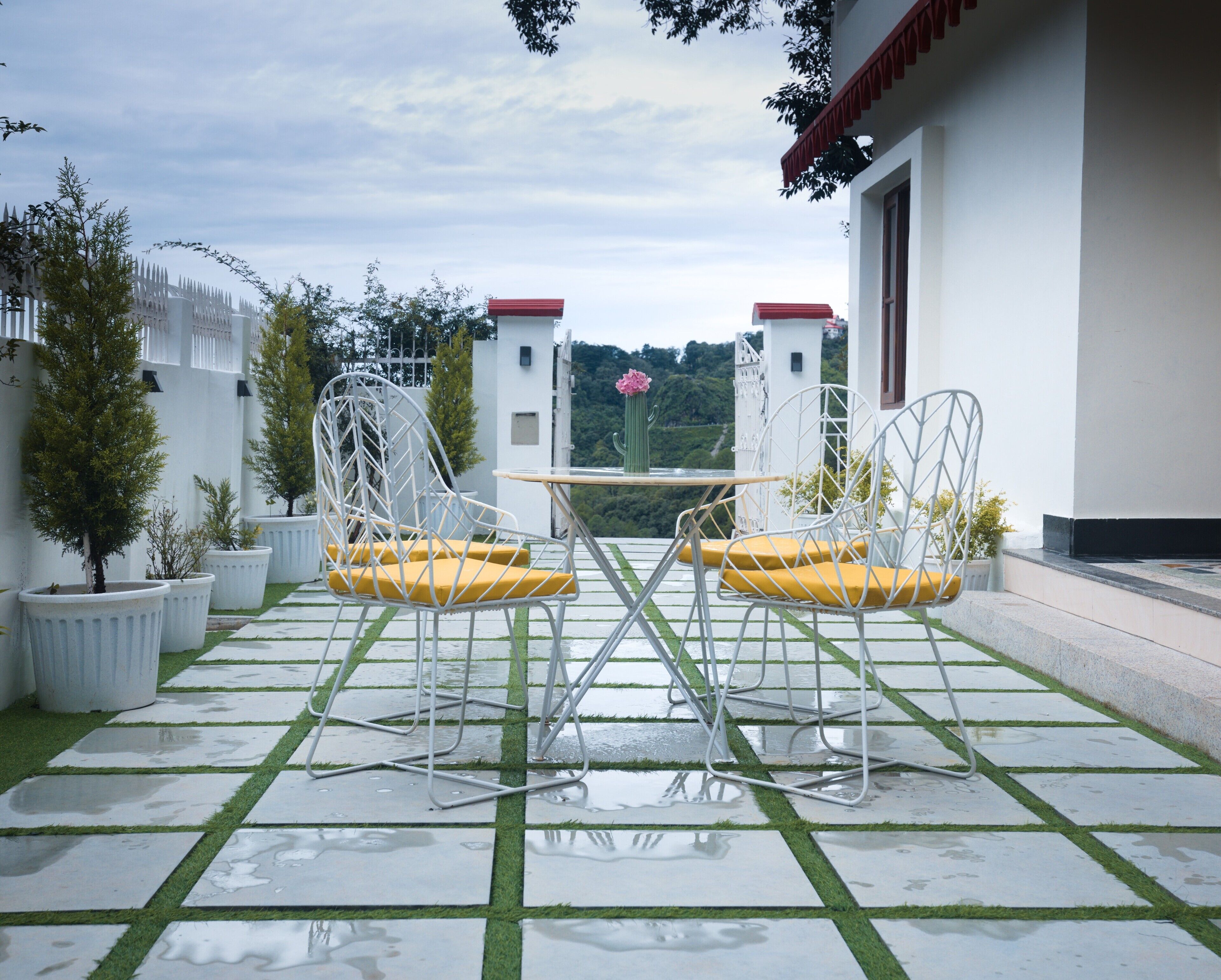 A patio with two white chairs and a table, surrounded by potted plants and a white building with red roofs.