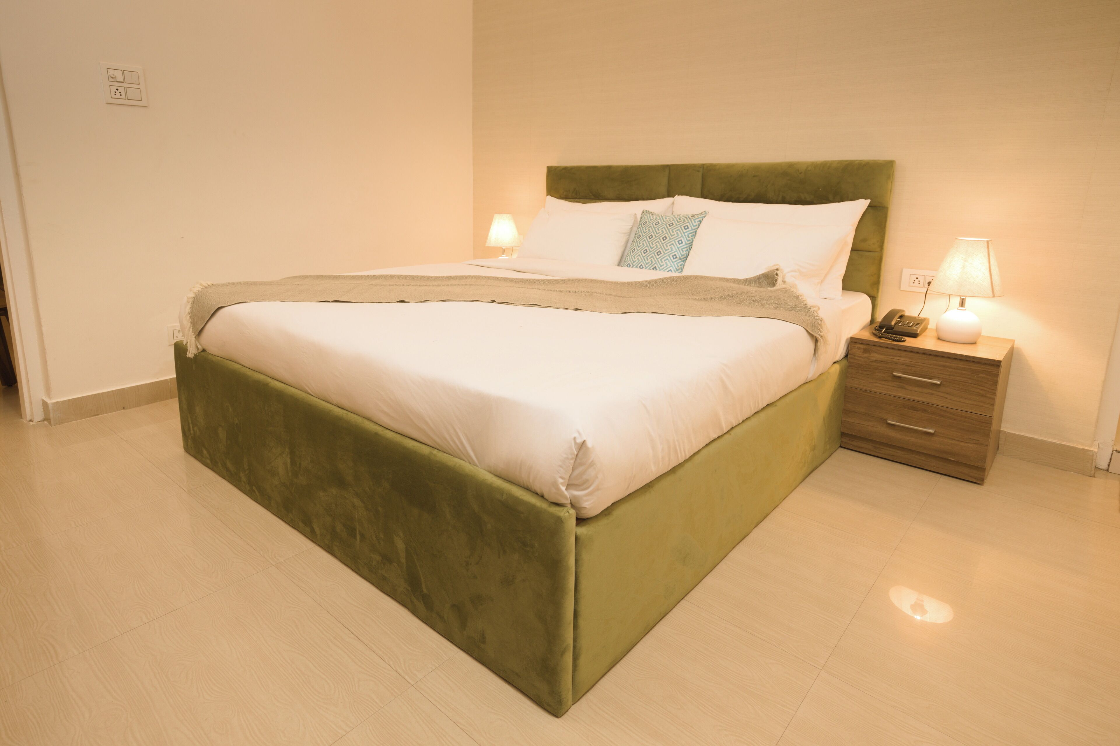 A neatly made bed with a green headboard, a bedside table with a lamp, and a drawer unit.