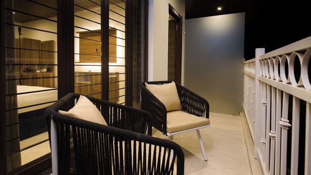 Un balcón con dos sillas de mimbre negro con cojines color beige, baranda blanca y vista a una habitación con cama y mesita de noche.