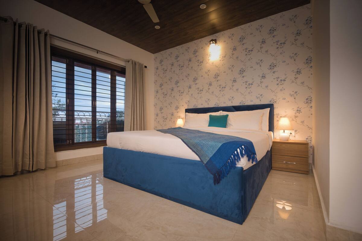 Ein Schlafzimmer mit einem großen Bett, einem Nachttisch, einem Fenster mit Vorhängen und einer Wand mit Blumenmuster-Tapete.